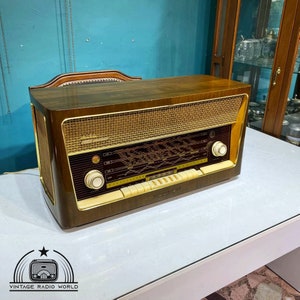 May include: A vintage wooden radio with a gold-colored front panel and a black dial. The radio has a wood case and a gold-colored grille. The radio is sitting on a white table.