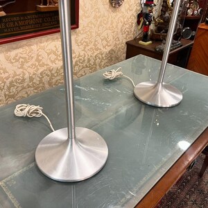 May include: Two silver table lamps with a modern design. The lamps have a tall, slender base and a wide, circular shade. The lamps are sitting on a wooden table with a glass top.