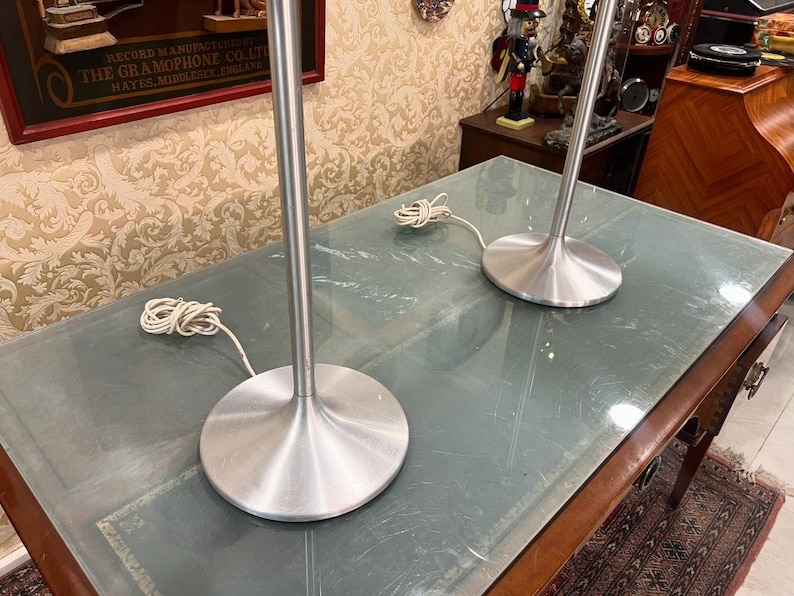 May include: Two silver table lamps with a modern design. The lamps have a tall, slender base and a wide, circular shade. The lamps are sitting on a wooden table with a glass top.