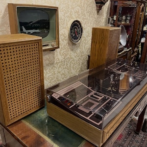 Puede incluir: Un conjunto de entretenimiento vintage con un televisor retro que muestra un avión, dos altavoces de madera con frentes tejidos y un tocadiscos bajo una cubierta transparente. Un reloj está montado en la pared.
