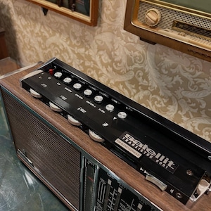 May include: A vintage radio with a wood grain finish and a black control panel. The radio has a silver dial and the text "Satellit Transistor 6001 SW-BAND SPREAD". The radio is on a green surface.