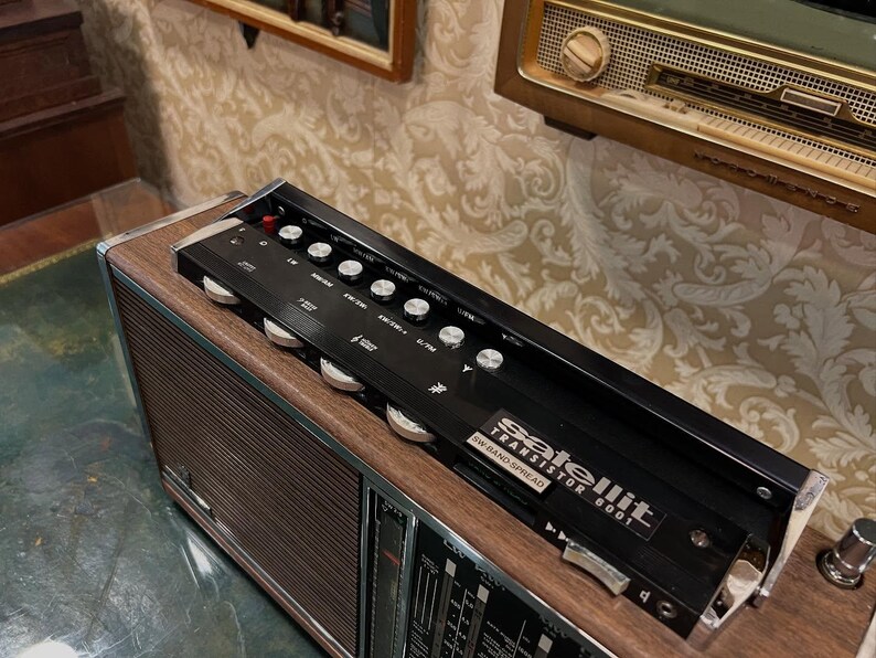 May include: A vintage radio with a wood grain finish and a black control panel. The radio has a silver dial and the text "Satellit Transistor 6001 SW-BAND SPREAD". The radio is on a green surface.