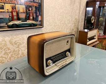 Puede incluir: Una radio vintage de madera con acabado marrón y crema. La radio tiene una rejilla de altavoz con un patrón tejido y un dial con botones de sintonización. La radio está sobre una mesa de vidrio.