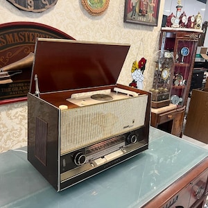 Puede incluir: Un tocadiscos vintage de madera con una tapa marrón abierta, que revela una plataforma giratoria y una rejilla de altavoz de color crema. El tocadiscos tiene un borde negro y dorado y una perilla plateada.