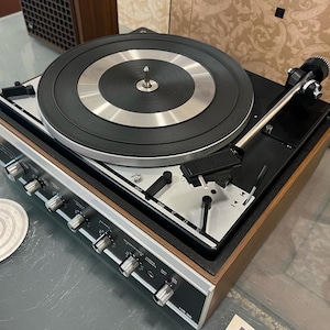 Może przedstawiać: Winylowy gramofon vintage z drewnianym korpusem i srebrnymi akcentami. Gramofon ma czarny talerz i srebrny tonearm. Na przednim panelu gramofonu znajduje się kilka pokręteł, w tym pokrętła głośności, tonu i balansu.