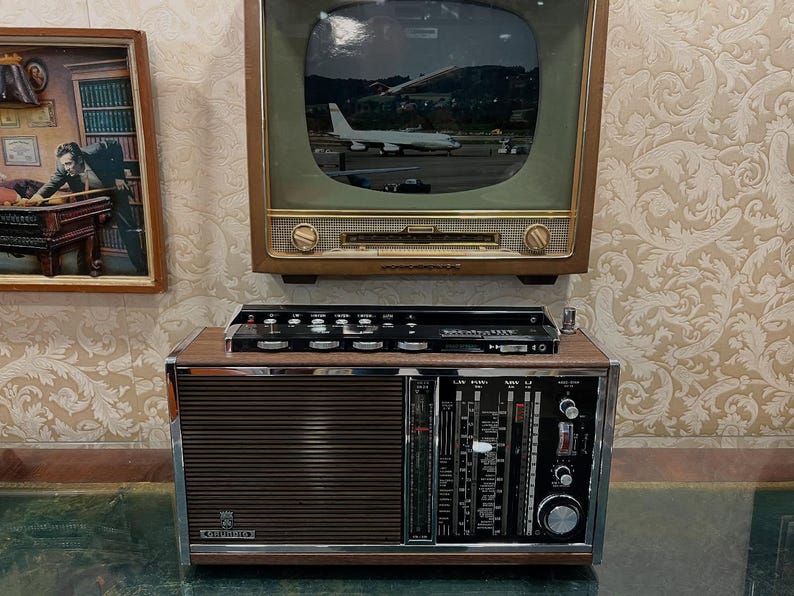 May include: Vintage radio with a wood and metal finish, featuring a speaker grill and control panel. Above it is a vintage television displaying an image of an airplane. A framed picture hangs on the wall.