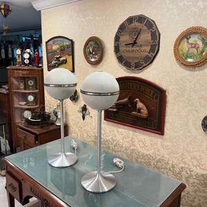 May include: Two vintage, white, globe-shaped speakers with silver accents and metal stands. The speakers are on a wooden table with a glass top. The speakers are in front of a wall with various decorative items, including a clock, a framed print, and a framed picture.