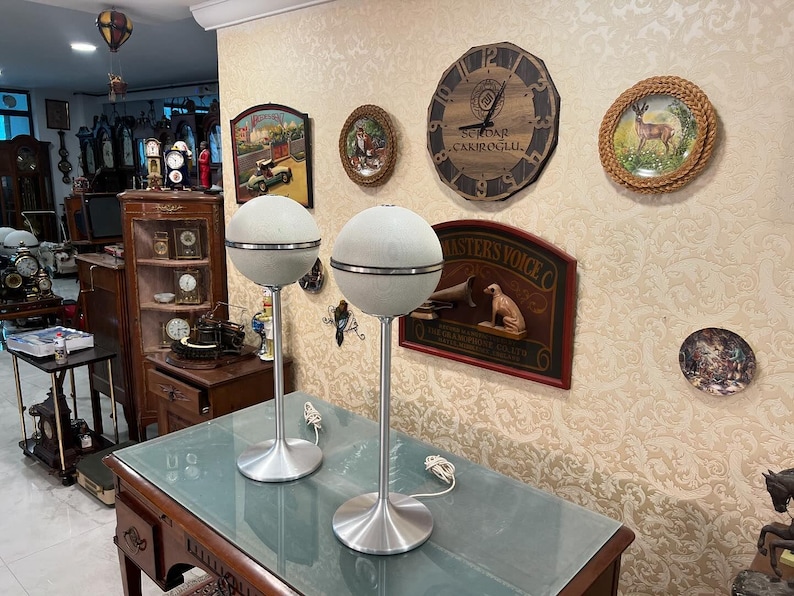 May include: Two vintage, white, globe-shaped speakers with silver accents and metal stands. The speakers are on a wooden table with a glass top. The speakers are in front of a wall with various decorative items, including a clock, a framed print, and a framed picture.