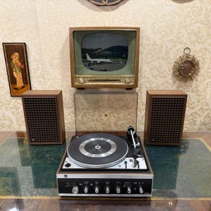 May include: A vintage stereo system featuring a turntable, two speakers, and a small television. The turntable is black and silver, with a clear cover. The speakers and TV have wooden frames. The TV screen displays an image of an aeroplane.