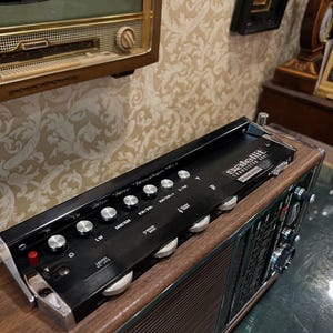 May include: A vintage black and wood-tone radio with silver knobs and the word "satellit" printed on the top. The radio has a retro design and is likely from the mid-20th century. The radio is sitting on a wooden surface.
