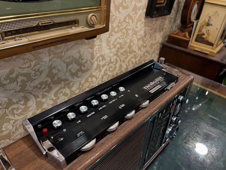 May include: A vintage black and wood-tone radio with silver knobs and the word "satellit" printed on the top. The radio has a retro design and is likely from the mid-20th century. The radio is sitting on a wooden surface.