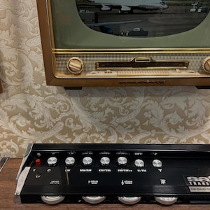 May include: Vintage black Satellit transistor radio with silver knobs and the text "Satellit Transistor S001 SW Band Spread." Above it is a vintage television with a gold frame. The background is a patterned wallpaper.