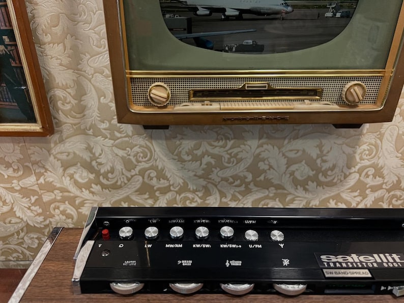 May include: Vintage black Satellit transistor radio with silver knobs and the text "Satellit Transistor S001 SW Band Spread." Above it is a vintage television with a gold frame. The background is a patterned wallpaper.