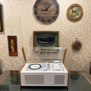 Puede incluir: Un tocadiscos vintage blanco y con detalles en madera con una cubierta de plástico transparente. El tocadiscos tiene un plato giratorio, un panel de control y rejillas de altavoz. Un televisor y un reloj vintage están montados en la pared.