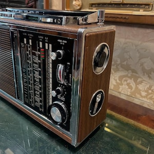 May include: A vintage radio with a wood grain finish and chrome accents. The radio features a tuning dial, various knobs, and a speaker grill. The face of the radio has a black panel with white and red markings. The radio is a retro style.