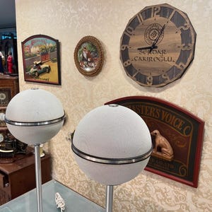 May include: Two white spherical speakers with black trim on stands. The speakers are in front of a wall with various decorative items, including a clock with the text "SEVDAR &Ccedil;AKIROĞLU."