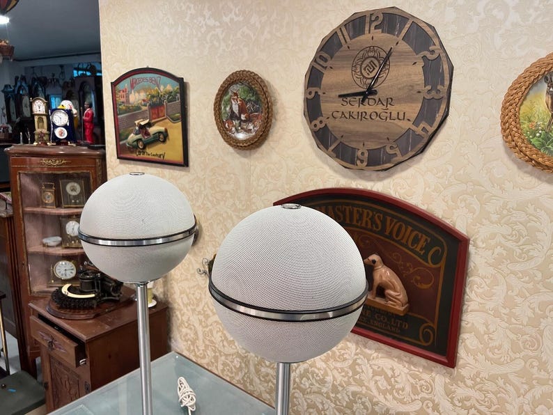 May include: Two white spherical speakers with black trim on stands. The speakers are in front of a wall with various decorative items, including a clock with the text "SEVDAR &Ccedil;AKIROĞLU."