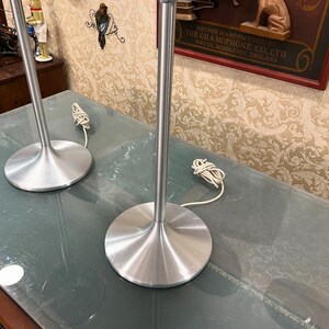 May include: Two silver table lamps with white cords on a glass table top. The lamps have a simple, modern design with a wide base and a tall, slender stem.