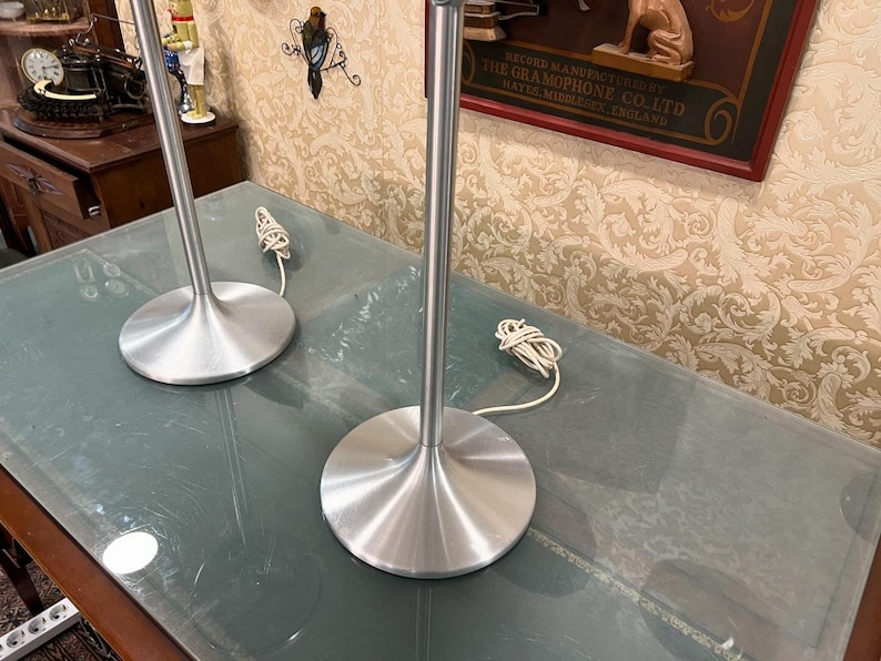 May include: Two silver table lamps with white cords on a glass table top. The lamps have a simple, modern design with a wide base and a tall, slender stem.