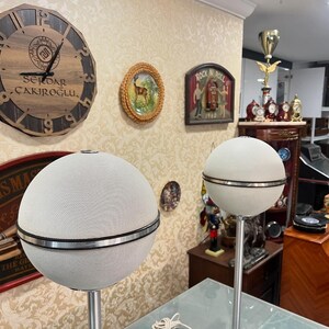 May include: Two white spherical speakers with a silver band around the middle, mounted on tall silver stands. The speakers are in a room with a wooden clock on the wall, a framed picture of a deer, and a framed picture of a rock and roll scene.