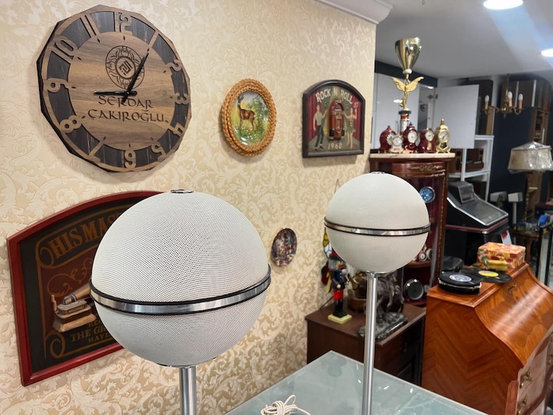 May include: Two white spherical speakers with a silver band around the middle, mounted on tall silver stands. The speakers are in a room with a wooden clock on the wall, a framed picture of a deer, and a framed picture of a rock and roll scene.
