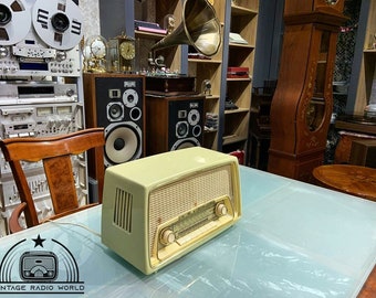 Puede incluir: Una radio vintage de color crema con un dial dorado y una rejilla de altavoz. La radio está sentada en una mesa de vidrio en una habitación con otros artículos vintage.