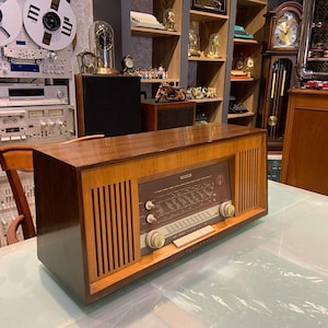 Puede incluir: Una radio vintage de madera con acabado marrón y dorado. La radio tiene un dial con una variedad de botones y perillas. La radio está sentada sobre una mesa blanca.
