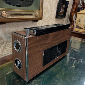 May include: Vintage wooden radio with chrome accents. The radio has a dark wood grain finish, a black top, and circular speaker covers. A rectangular vent is visible on the front. The radio is a retro style electronic device.