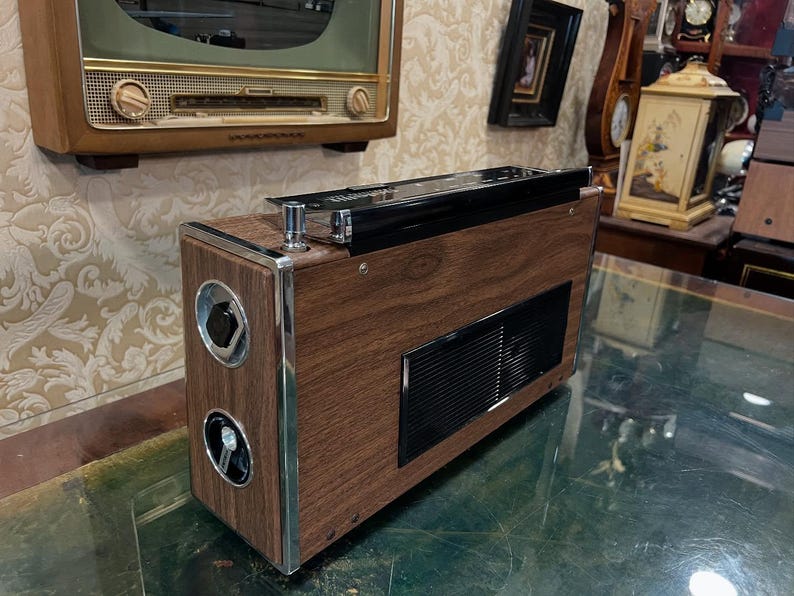 May include: Vintage wooden radio with chrome accents. The radio has a dark wood grain finish, a black top, and circular speaker covers. A rectangular vent is visible on the front. The radio is a retro style electronic device.