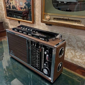 May include: Vintage wooden radio with a black top panel and silver accents. The radio has a brown speaker grill and a control panel with dials and gauges. The radio is sitting on a reflective surface.