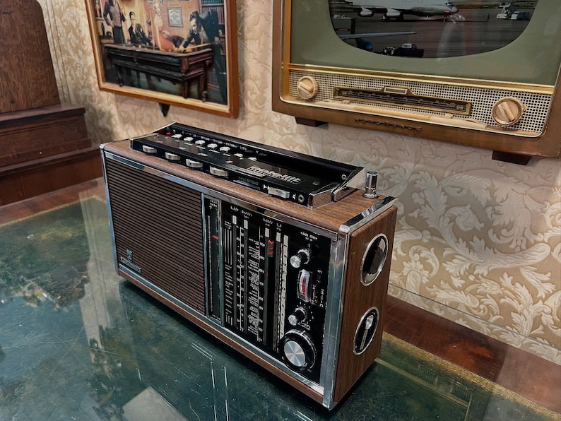 May include: Vintage wooden radio with a black top panel and silver accents. The radio has a brown speaker grill and a control panel with dials and gauges. The radio is sitting on a reflective surface.