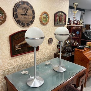 May include: Two white vintage mid-century modern speakers with a silver trim on a glass table top. The speakers are on a stand with a silver base.