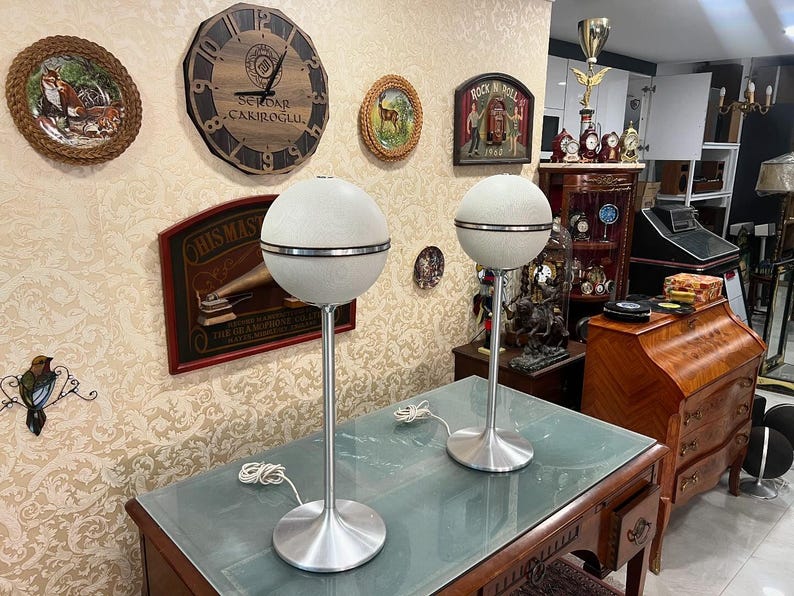 May include: Two white vintage mid-century modern speakers with a silver trim on a glass table top. The speakers are on a stand with a silver base.