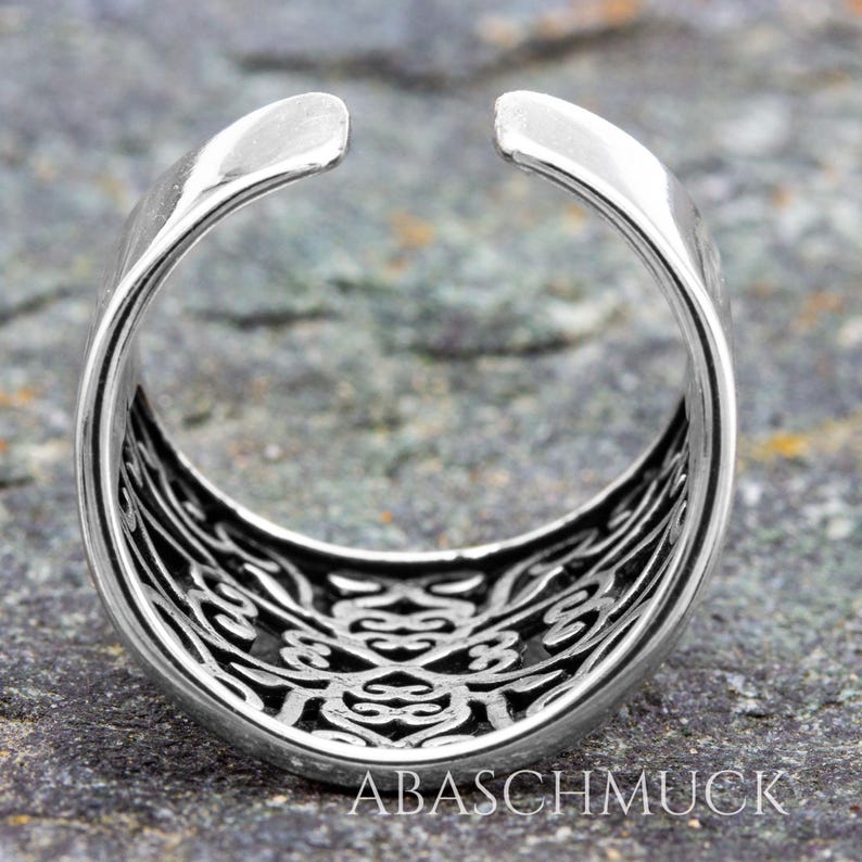 May include: A silver-colored ring with an open design. The ring features an intricate, openwork pattern on the inner band. The text "ABASCHMUCK" is visible at the bottom of the image. The ring is set against a textured, gray surface.