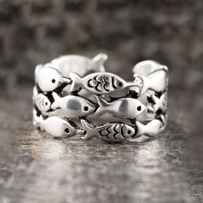 May include: Silver-toned ring featuring a repeating fish design. The ring has three rows of fish, each with detailed scales and eye markings. The ring is a wrap-around style, with the fish facing in different directions.