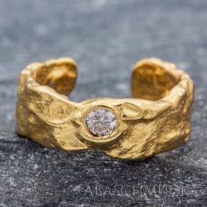 May include: A gold ring with a textured surface and a single clear gemstone in the centre.
