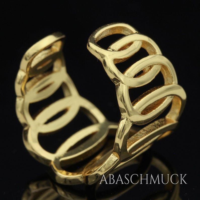 May include: A gold-colored ring with a chain-link design. The ring is made of a shiny, reflective metal and features a series of interconnected oval shapes. The ring is displayed against a dark background. The text "ABASCHMUCK" is visible.
