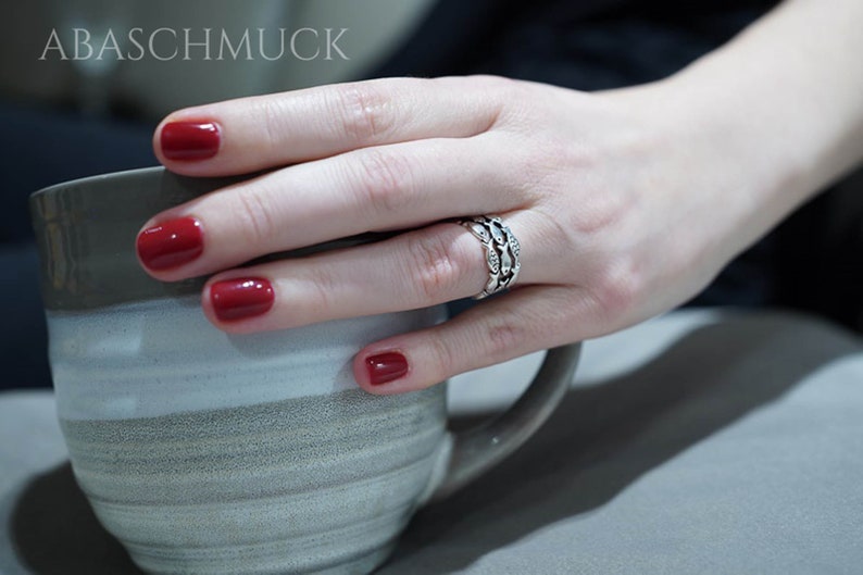 May include: A hand with red nail polish holds a gray and white mug. A silver ring with a detailed design is on the ring finger. The image has the text "ABASCHMUCK" at the top.
