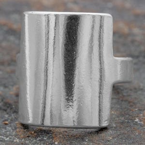 May include: A silver ring with a wide, rectangular band. The band has a textured, vertical pattern.