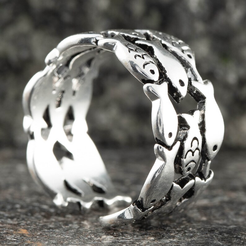 May include: A silver ring featuring a repeating pattern of fish. The ring has a detailed design with each fish individually crafted. The ring is made of a shiny, metallic material and is set against a dark background.