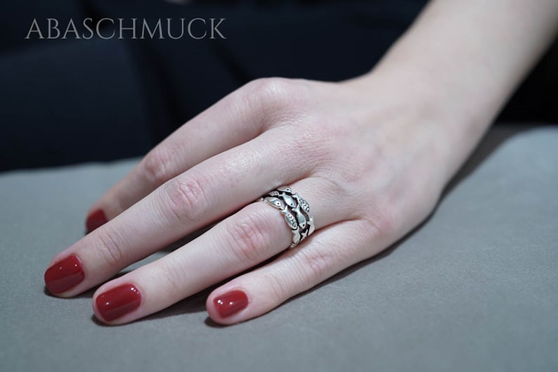 May include: A silver ring with a repeating leaf design, set with small clear stones. The ring is worn on a finger with red nail polish. The brand name "ABASCHMUCK" is visible in the background.