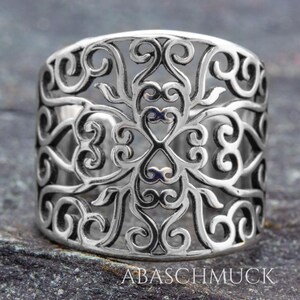 May include: A silver-colored ring with an intricate openwork design. The ring features a repeating pattern of swirling lines and heart-shaped elements. The brand name "ABASCHMUCK" is visible at the bottom of the ring.
