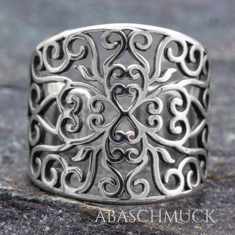 May include: A silver-colored ring with an intricate openwork design. The ring features a repeating pattern of swirling lines and heart-shaped elements. The brand name "ABASCHMUCK" is visible at the bottom of the ring.