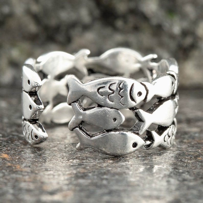 May include: A silver-toned ring featuring a repeating pattern of fish. The ring has a three-dimensional design with the fish arranged in rows around the band. The fish have detailed scales and eye markings. The ring is displayed on a textured gray surface.