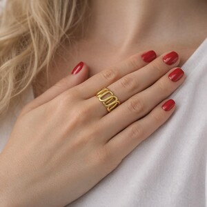 Peut inclure: Une bague dorée avec un design ovale ouvert. La bague est portée au doigt, la main reposant sur une chemise blanche. Les ongles sont peints en rouge. Le design de la bague présente une série d'ovales allongés et connectés.