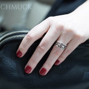 May include: A silver ring with a fish scale design is worn on a finger. The ring is on a hand with red nail polish. A black leather bag is in the background. The text "ABASCHMUCK" is at the top.