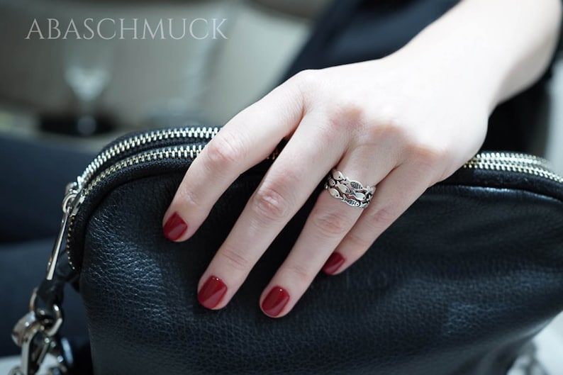 May include: A silver ring with a fish scale design is worn on a finger. The ring is on a hand with red nail polish. A black leather bag is in the background. The text "ABASCHMUCK" is at the top.