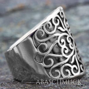 May include: A silver-colored ring with an openwork design featuring swirling patterns. The ring has a wide band and a decorative, curved top. The text "ABASCHMUCK" is visible on the ring.