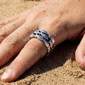 May include: A silver ring featuring a school of fish design. The ring is worn on a finger, with the details of the fish visible. The ring is set against a sandy background, suggesting a beach or coastal theme.