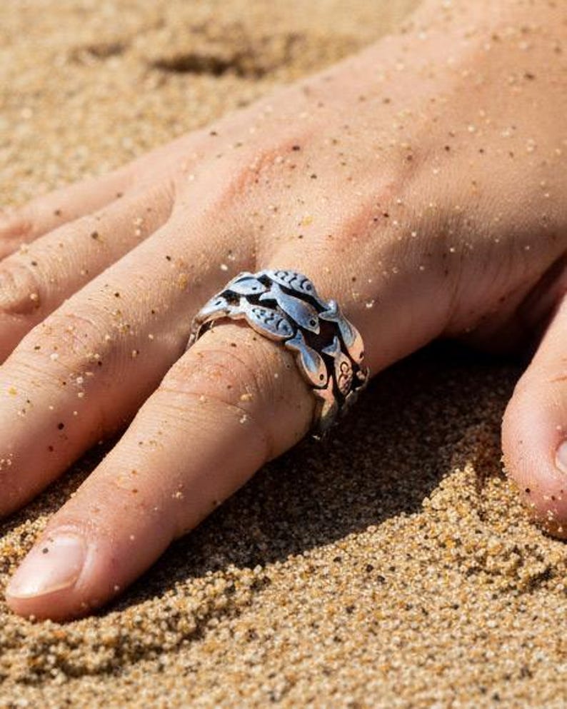May include: A silver ring featuring a school of fish design. The ring is worn on a finger, with the details of the fish visible. The ring is set against a sandy background, suggesting a beach or coastal theme.